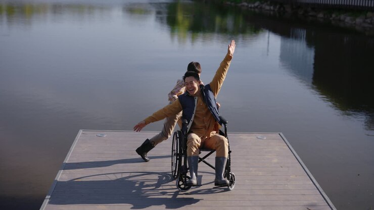 Boy Helping Man in Wheelchair on a Dock by a Lake