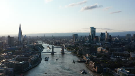 Aerial-panoramic-view-of-River-Thames-flowing-through-city.-Old-Tower-Bridge-contracting-with-modern-skyscrapers.-Scene-lit-by-evening-sun.-London,-UK