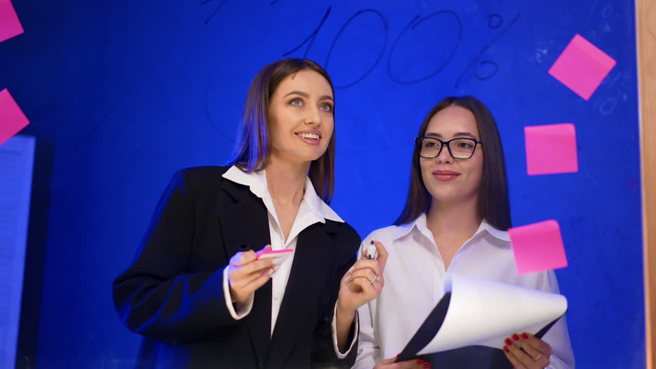Positive Caucasian ladies stand behind the glass wall. Woman in jacket talks to colleague and writes on the board.