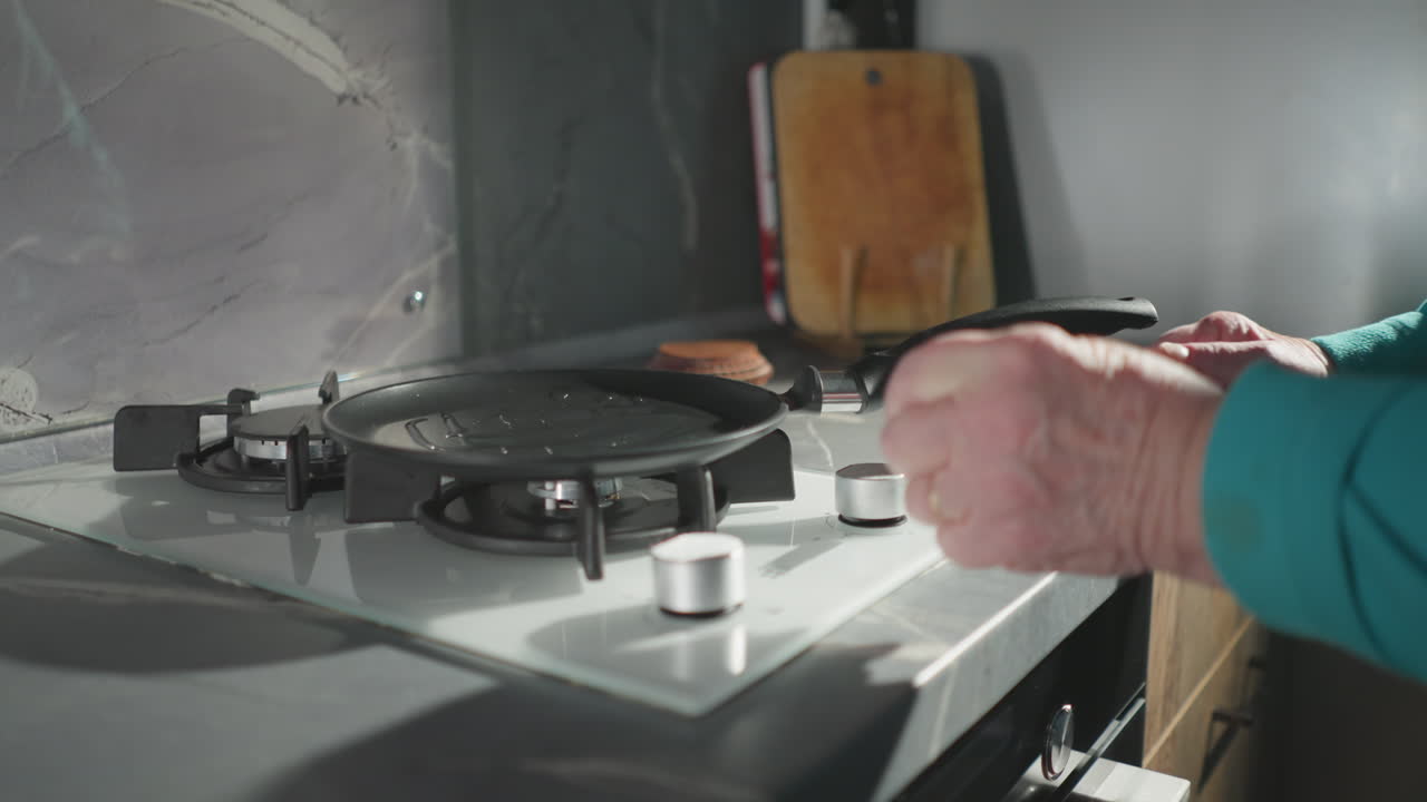 Elderly woman in green sweatshirt using pan on stove in modern kitchen. Focused on preparing meal, demonstrating independence and self-sufficiency. Modern kitchen with sleek surfaces and natural light