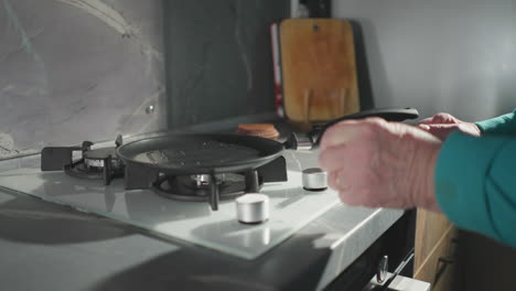 elderly woman in green sweatshirt using pan on stove in modern kitchen. focused on preparing meal, demonstrating independence and self-sufficiency. modern kitchen with sleek surfaces and natural light