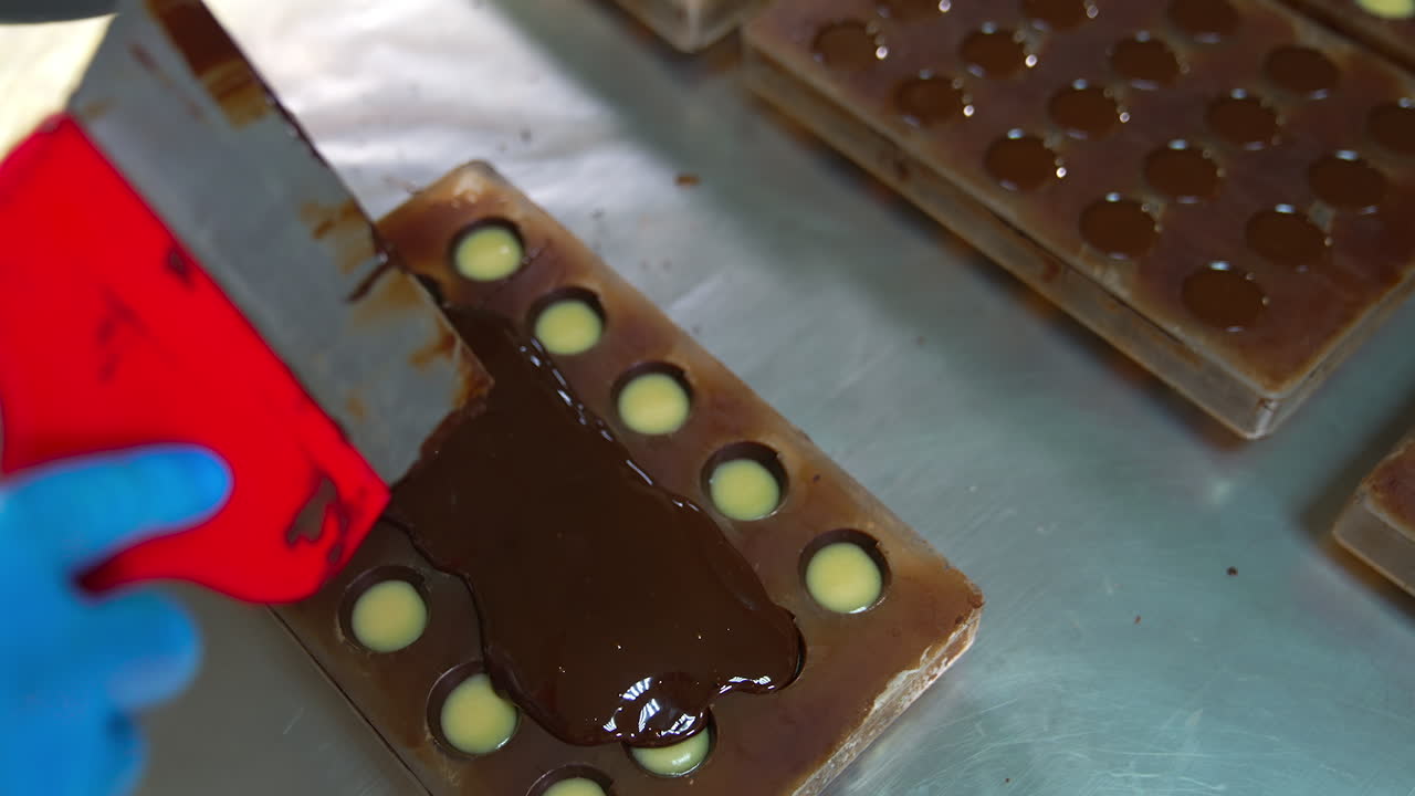 Chocolatier pours black melted chocolate on the filled molds. Gloved hand spreads the chocolate with big spatula. Close up.
