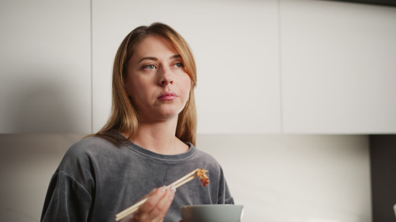 Lady eating meat with chopsticks inside bright modern kitchen, drops meat into bowl then picks another piece, standing against clean white cabinetry, dressed casually, enjoying her meal