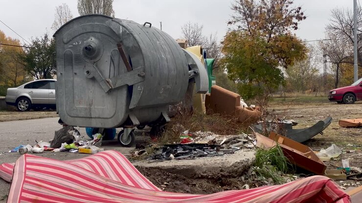 Bidone della spazzatura straripante e sacchi di rifiuti di plastica che causano un problema ambientale, divano sullo sfondo.