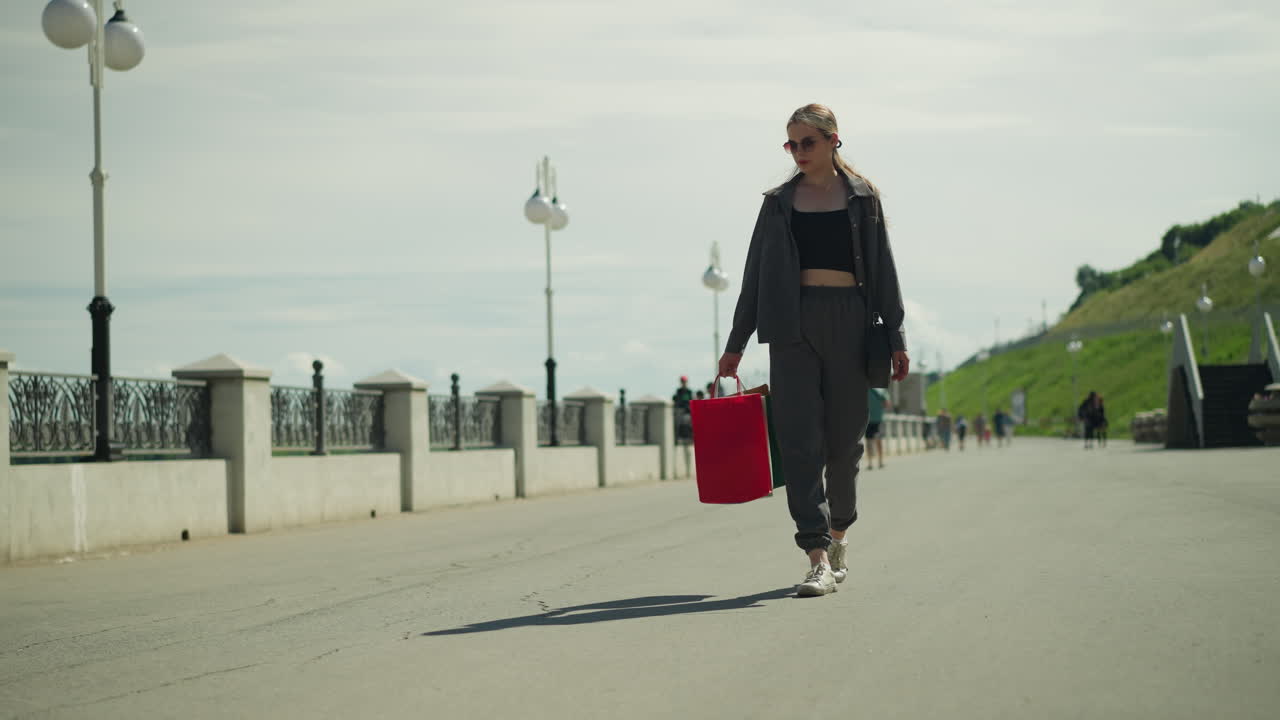 Lady wearing sunshades holding red, mint, and green shopping bags, looking down as she walks then glancing away, her shadow is seen on the tarred road, background is blur with other people