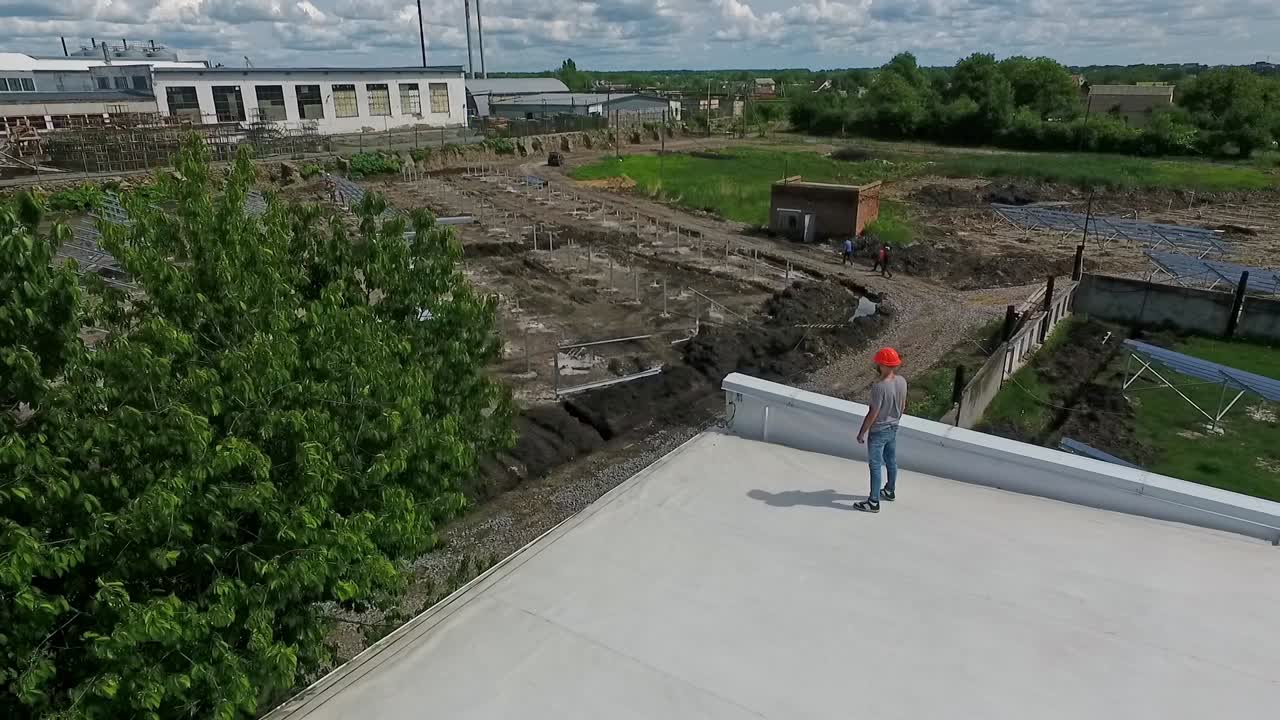 Technician in orange hard hat on a roof in summer. Installer looking on the solar farm construction from a rooftop of a building. Aerial view.