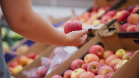 partial back view of woman picking a red apple from a box of apples, briefly observing it, and then returning it to the box while shopping at a grocery store or market