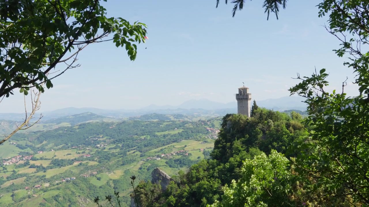 Una torre medievale in pietra si erge su un crinale boscoso mentre un parapendio sorvola valli ondulate e terreni agricoli a San Marino