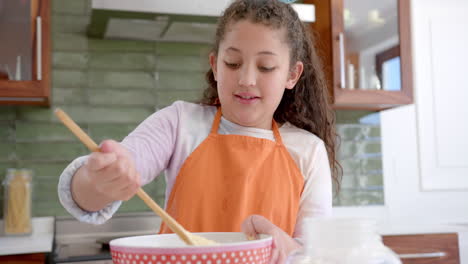 Happy-biracial-girl-with-long,-curly-hair-mixing-dough-in-bowl-and-smiling-in-sunny-kitchen