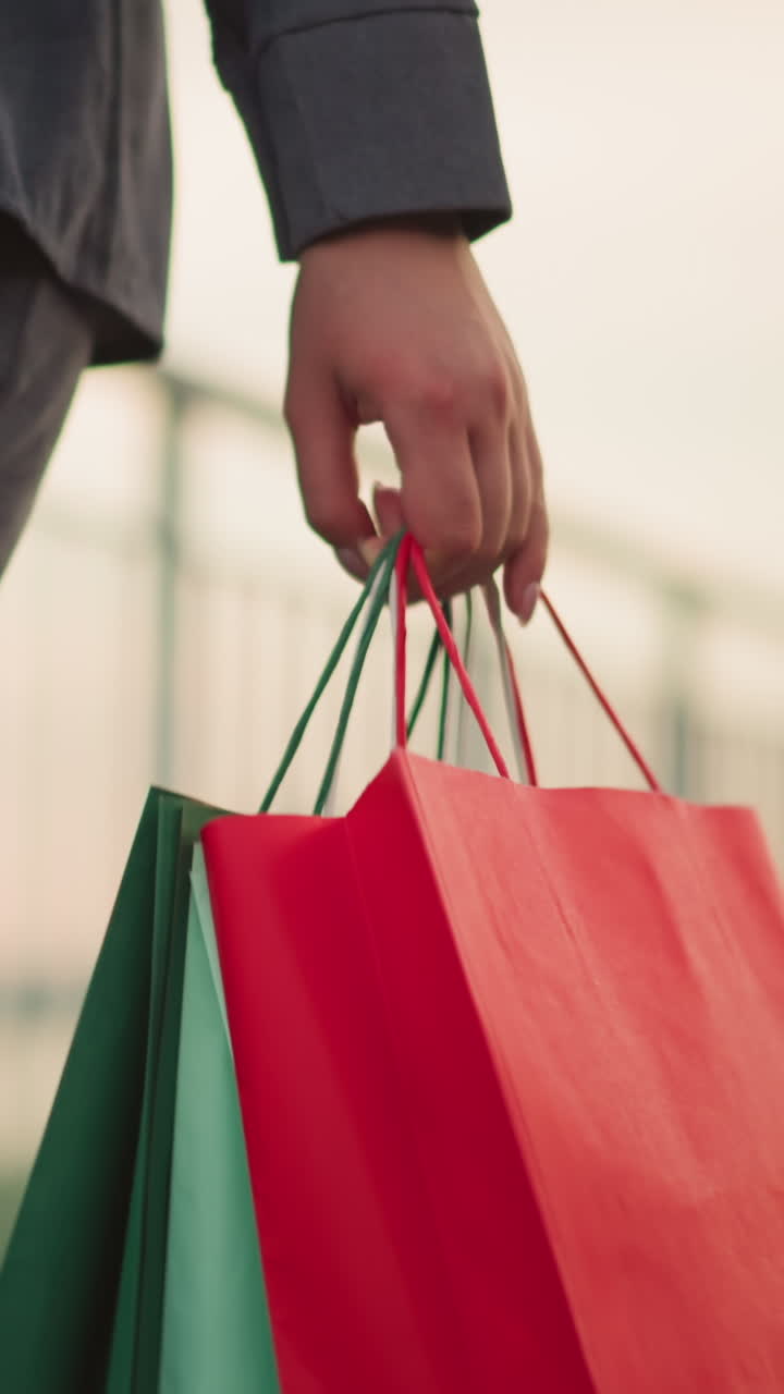 primer plano de un individuo con ropa gris que lleva tres bolsas de compras de colores mientras camina a lo largo de un carril de hierro, con un fondo borroso al amanecer