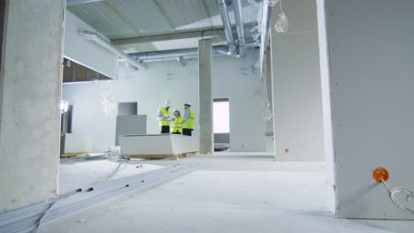 team of engineers in hard hats having conversation, looking at blueprint, inside building under construction.