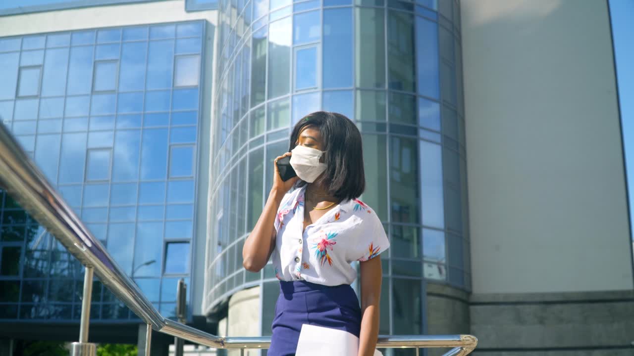 보호 마스크를 입은 성인 아프리카계 미국인 젊은 사업가 여성이 현대 사무실 건물 근처 밖에서 전화로 이야기하고 있습니다.