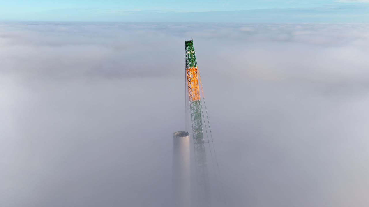 Aerial approach shot of a tall construction crane assembling a wind turbine, surrounded by thick clouds in a Latvian wind farm at sunrise