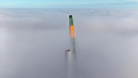 aerial approach shot of a tall construction crane assembling a wind turbine, surrounded by thick clouds in a latvian wind farm at sunrise