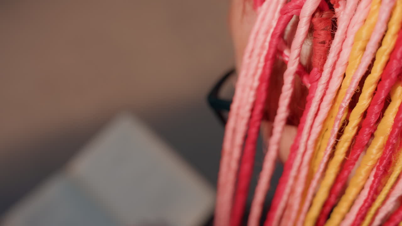 Primer plano de una textura de trenza multicolor con hebras rosas y amarillas, fibras detalladas y un libro suavemente desenfocado al fondo; detalle de moda que destaca el contraste de color, el estilo y el patrón de trenza artesanal