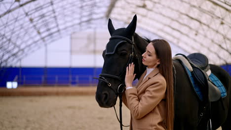 attractive woman with a black horse in the riding hall. long-haired lady caressing the animal and leaning to it. blurred backdrop.