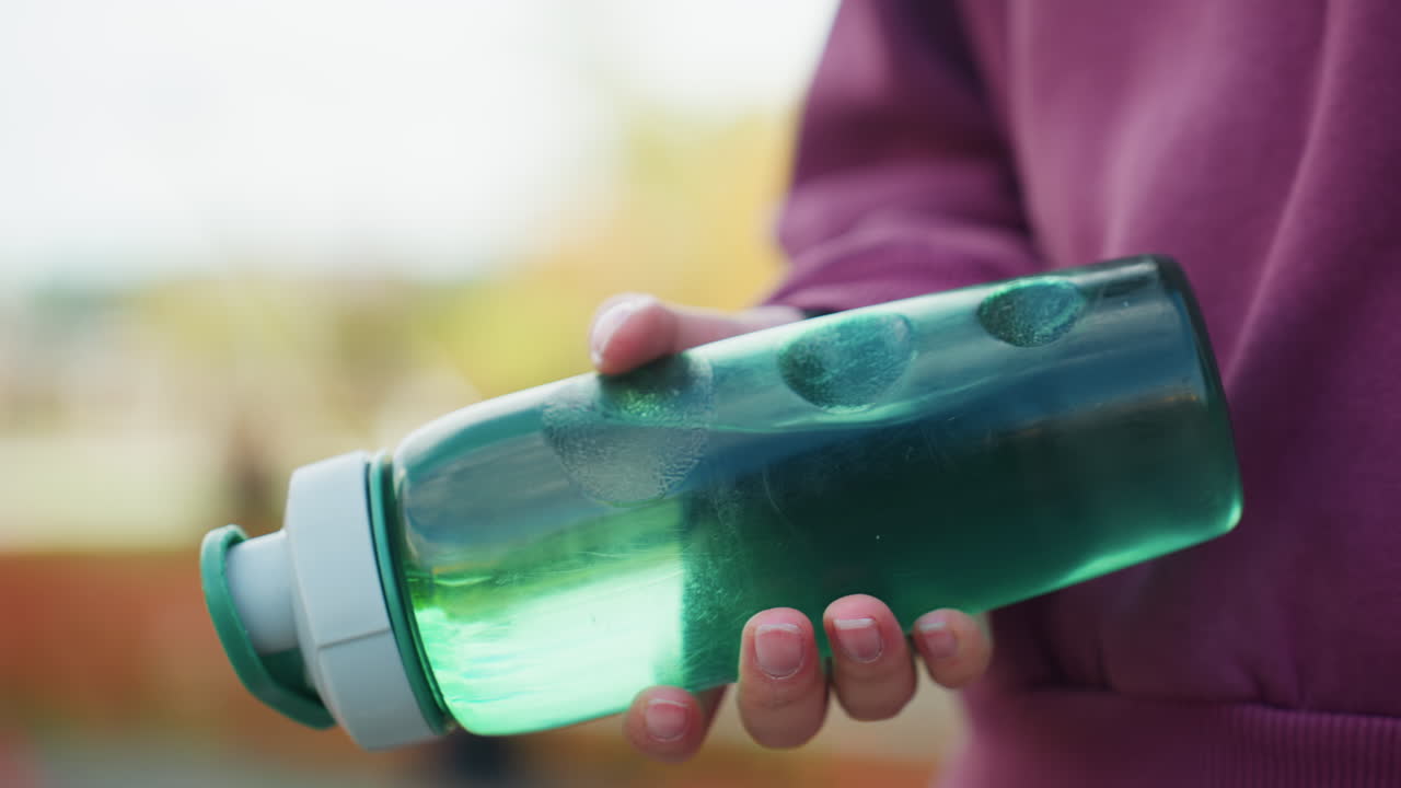 Close up city dweller hand gripping translucent green water bottle visible swirl of liquid while walking on urban pavement conveying dynamic hydration and fitness motion in outdoor scene