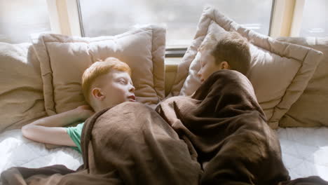 two little brothers talking and laughing together while lying under a blanket in the campervan bed