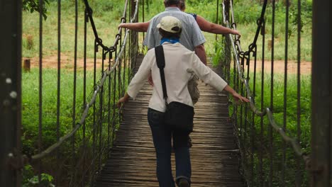 Three-adults-walk-over-swaying-suspension-bridge-made-of-reeds-and-steel-cables,-toward-grassy-green-wilderness-area-in-South-Africa