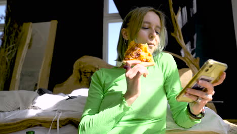 young blonde woman using mobile phone and eating pizza while sitting on the floor in the bedroom at home 2