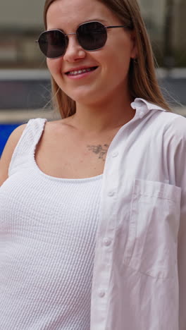 smiling blonde girl in sunglasses and white swimsuit and shirt enjoys cocktail. portrait of a happy lady resting at the pool. vertical video