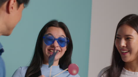 Group-Of-Young-Friends-In-Front-Of-Blue-Studio-Background-Having-Fun-Posing-For-Photo-Booth-Style-Portraits-With-Props-3