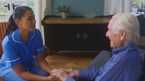 Female-Care-Worker-In-Uniform-Holding-Hands-Of-Senior-Man-Sitting-In-Care-Home-Lounge