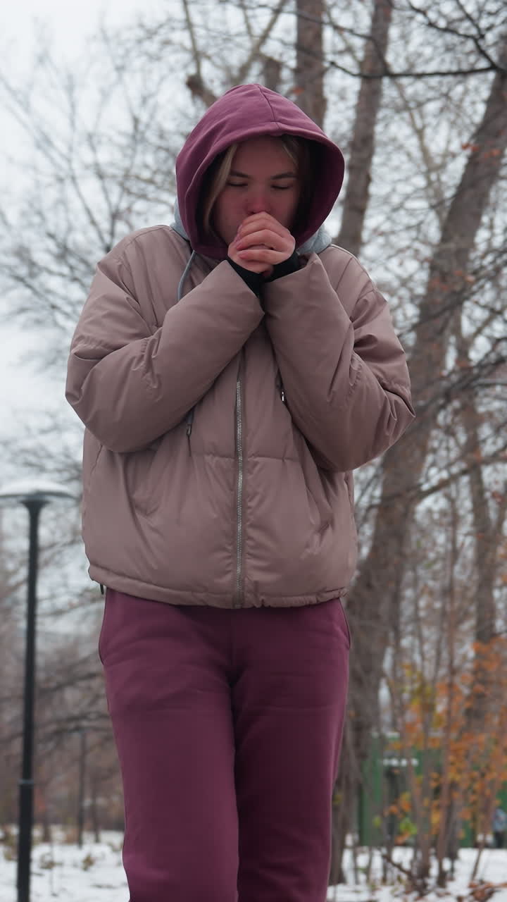 mujer joven vestida con ropa cálida de invierno camina a través de un parque nevado, sosteniendo sus manos cerca de su cara para el calor, árboles desnudos con bayas rojas, farolas distantes