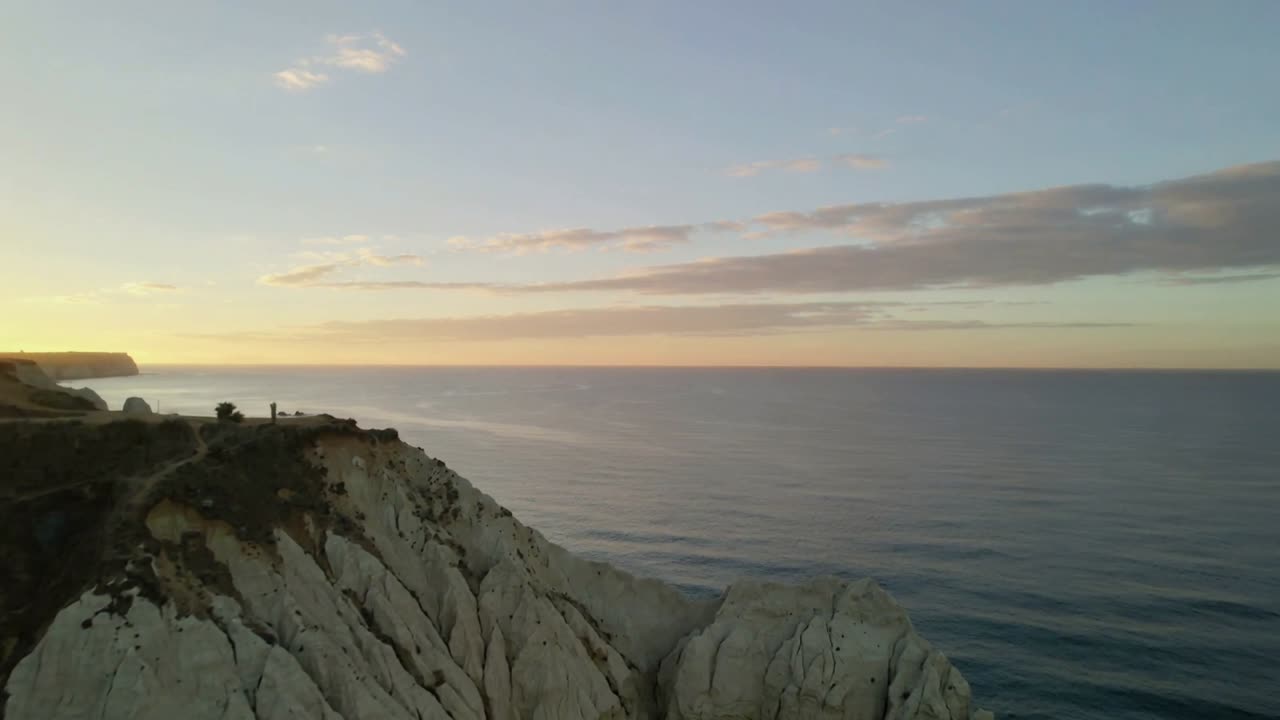 Dron panorámuje krémový útes se dvěma lidmi v tmavých kabátech, kteří kráčejí po okraji a pozorují západ slunce.