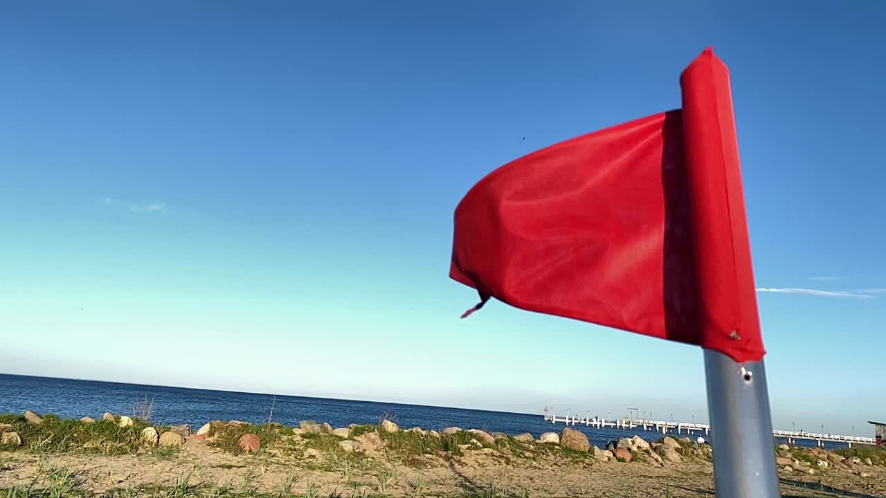 Bandera roja en blanco sobre un fondo del mar, símbolo de peligro, cielo azul sin nubes y muelle blanco a lo lejos.