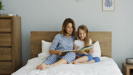 joven y hermosa madre acostada en la cama debajo de la manta y leyendo un libro con su hija por la mañana