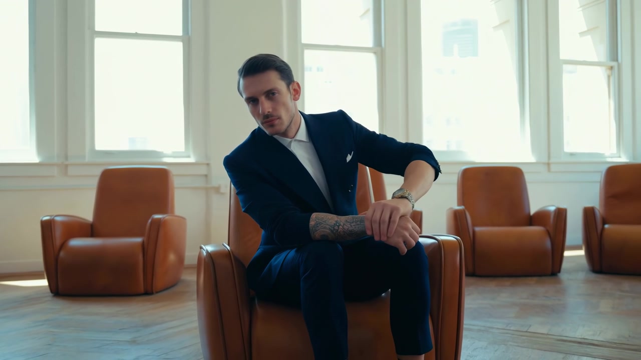 Giovane uomo in abito blu notte e camicia bianca siede al centro di una poltrona lounge in pelle color cuoio all'interno di una luminosa sala d'attesa minimalista, leggermente proteso in avanti, con le mani incrociate sul ginocchio e la postura leggermente ruotata.