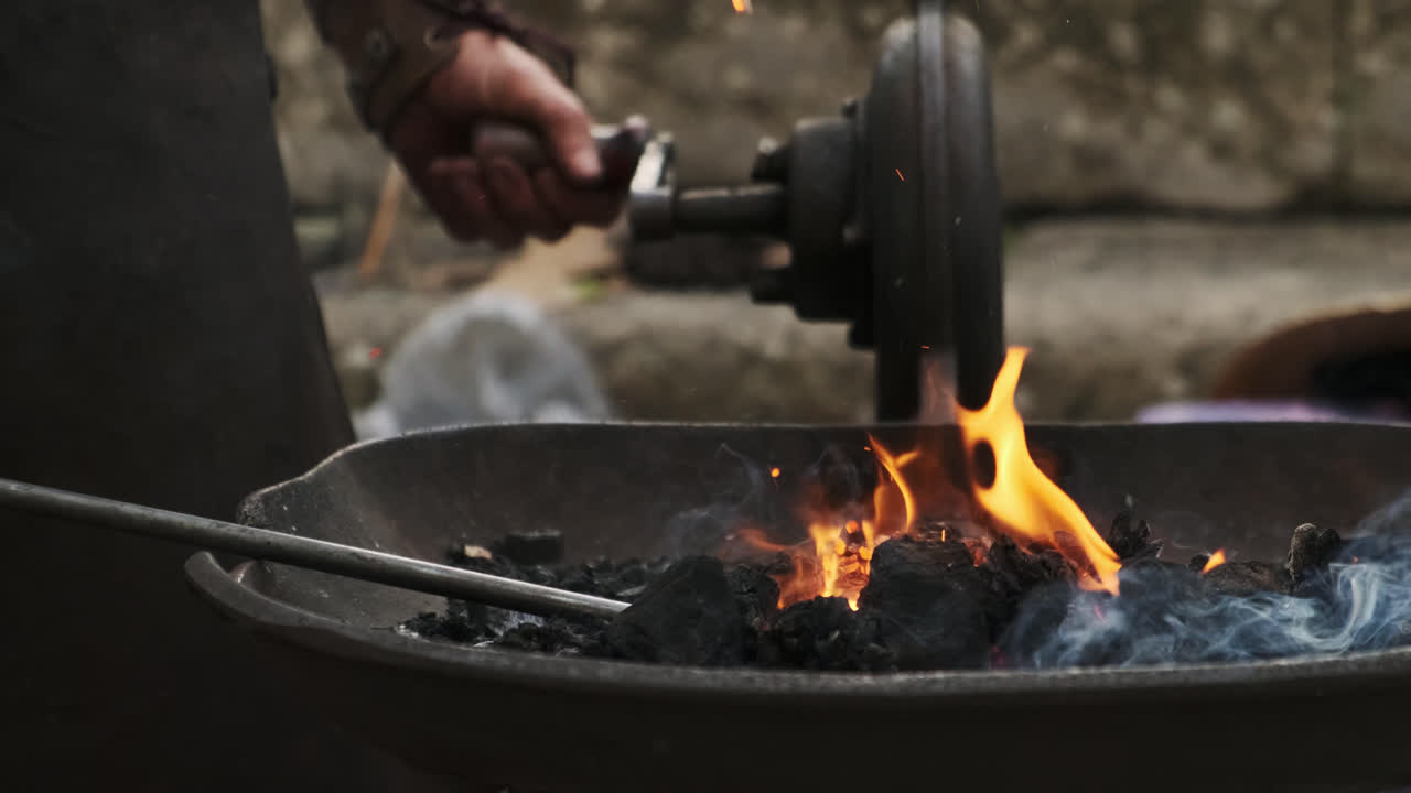 Frontal static close-up of a coal forge as a blacksmith uses a tool to rearrange hot coals; bright flames, flying sparks, and smoke rise from the brazier