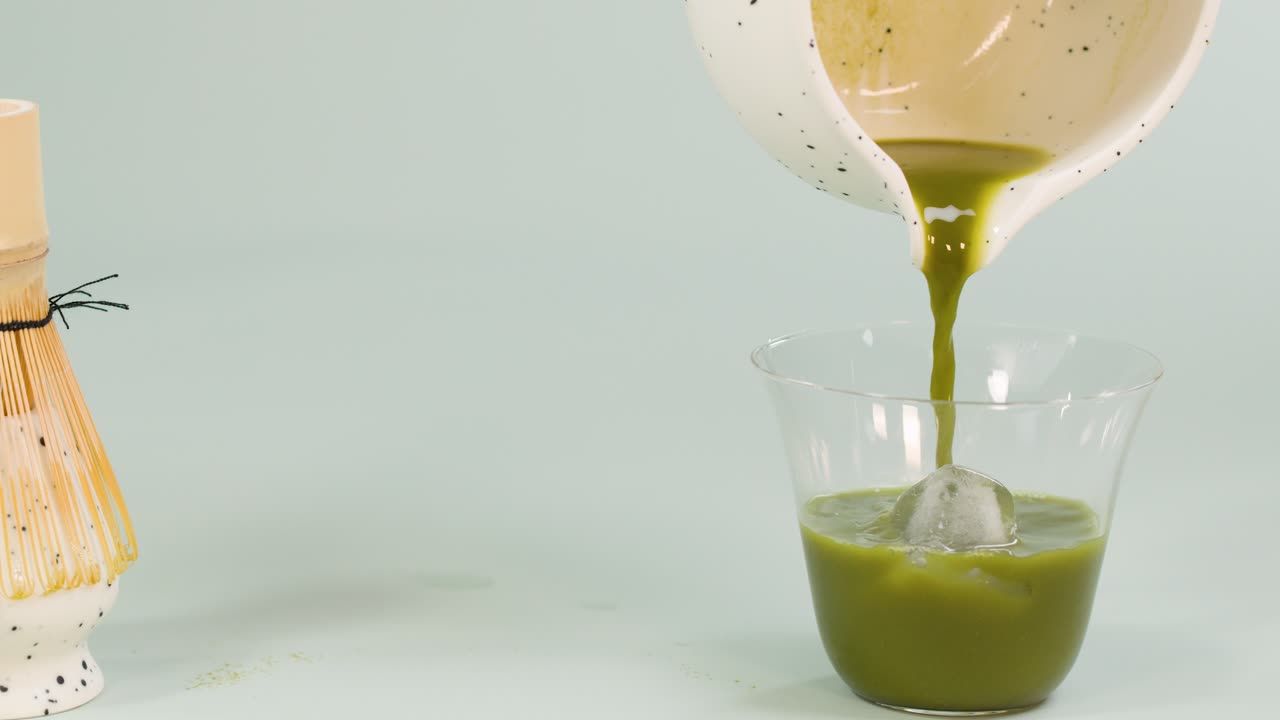 Hand pours vibrant matcha tea from ceramic bowl into glass with ice, soft studio lighting