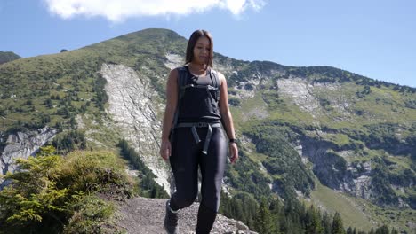 female hiker with backpack and hiking boots walks on a stony path in the blazing sun with mountain range in the background