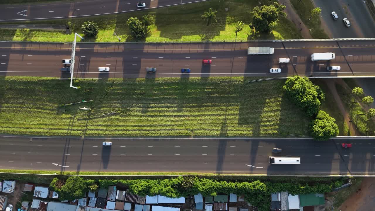 Drone clip of a busy highway scene as cars and trucks pass a poor community on the side of the road