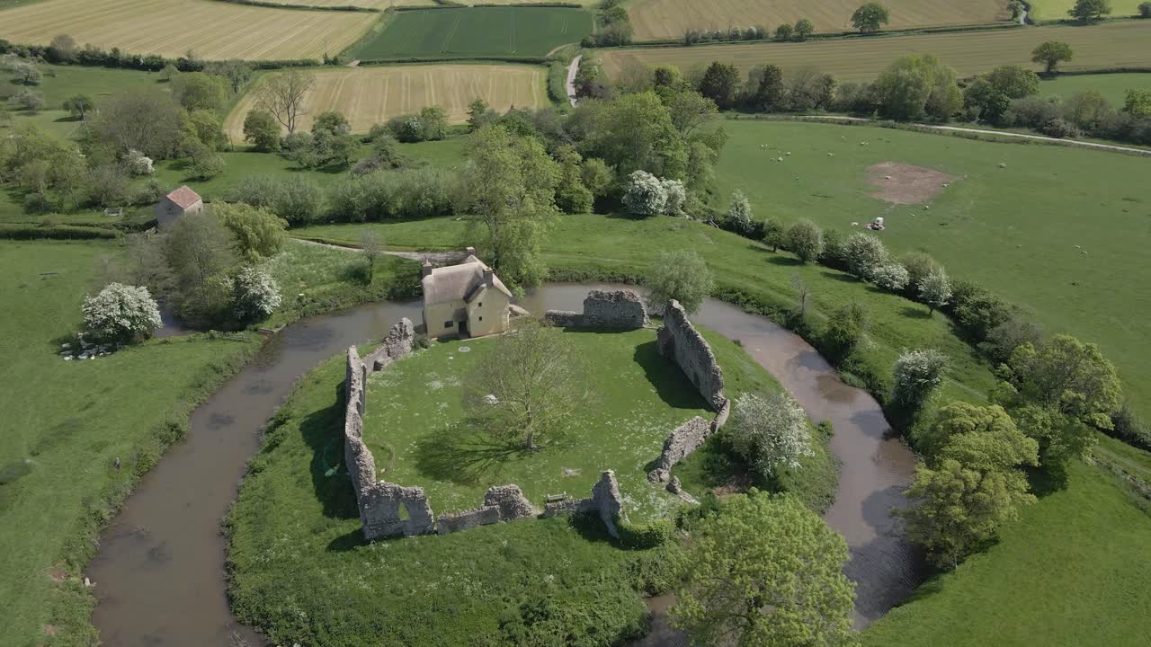 Aerial view of Stogursey Castle, a medieval castle in Somerset, England. Most of the site is in ruins, with a thatched gatehouse used for holiday's. Drone rotating to the left over the ruins and moat