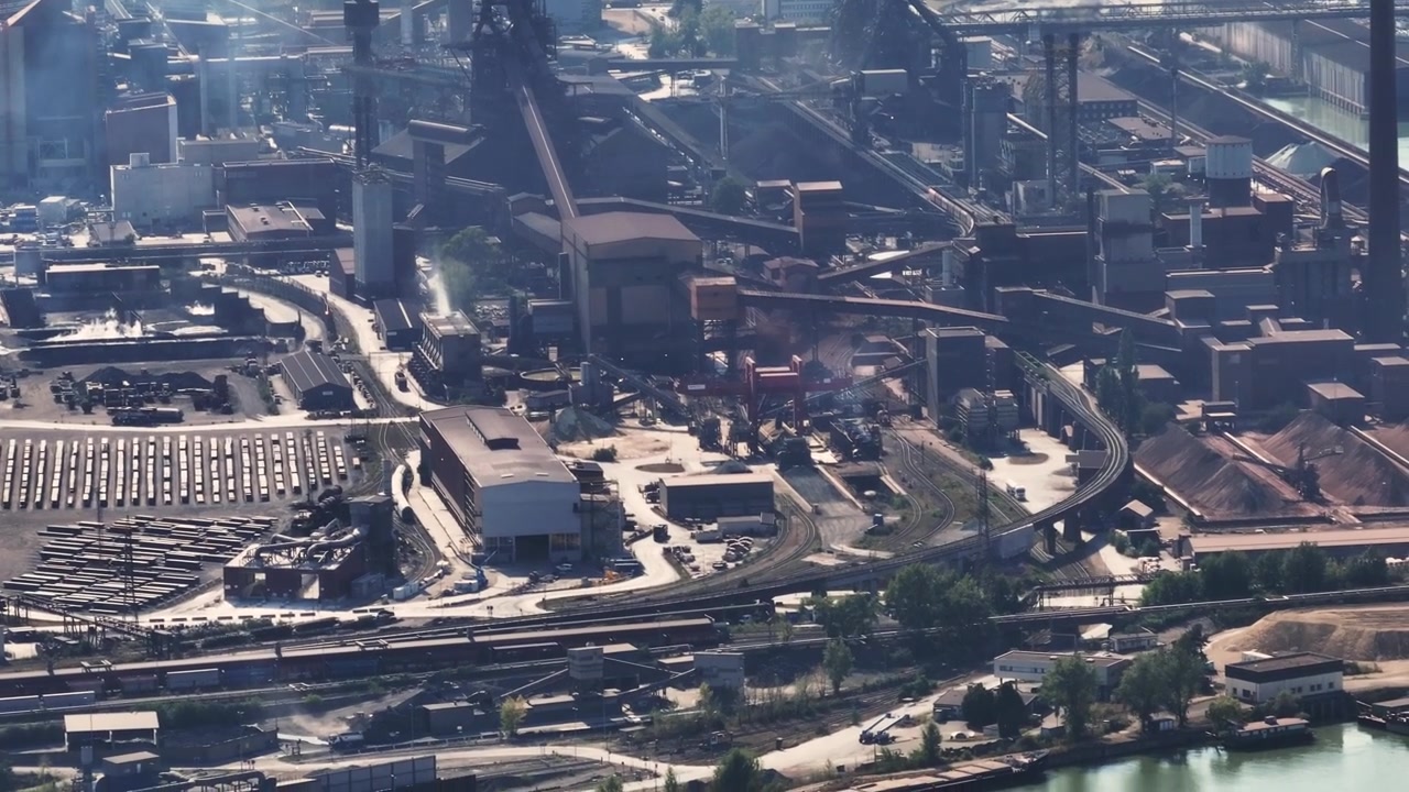 Aerial view of a large metallurgical complex with blast furnaces and chimneys producing smoke, located by a river and near a city