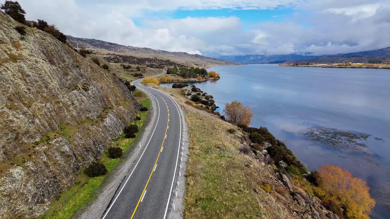 Drone glides above a winding lakeside road bordered by rocky hills and calm water, under bright daylight with soft clouds and natural reflections