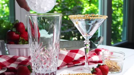strawberry shortcake martini cocktail with whipped cream, graham cracker crumbs and a fresh red strawberry. making a cocktail for happy hour. filling the crystal cocktail shaker with ice cubes.