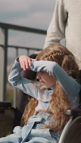 una niña interesada con una lesión en la pierna mira la ciudad cubriendo los ojos del sol brillante. madre empuja una silla de ruedas con su hija caminando por la acera cerca de la valla de primer plano