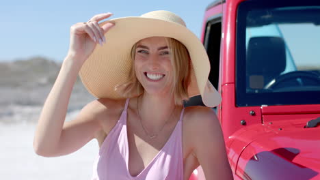 joven mujer caucásica sonríe sosteniendo su sombrero en la playa en un viaje por carretera