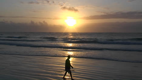 Un-Hombre-Camina-Por-La-Playa-Con-Un-Palo-Mientras-Las-Olas-Llegan-Y-El-Sol-Se-Pone