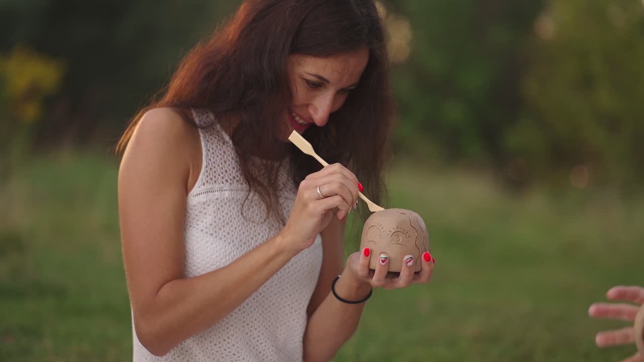 una joven mujer sonriente aplica un patrón un dibujo en un producto de arcilla con una herramienta un palo una pila en un prado en la naturaleza en un exterior. primer plano.
