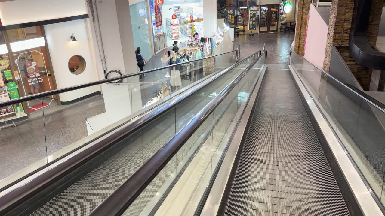 Smooth camera glide down escalator walkway, passing shops and bright retail lighting in Bangkok mall