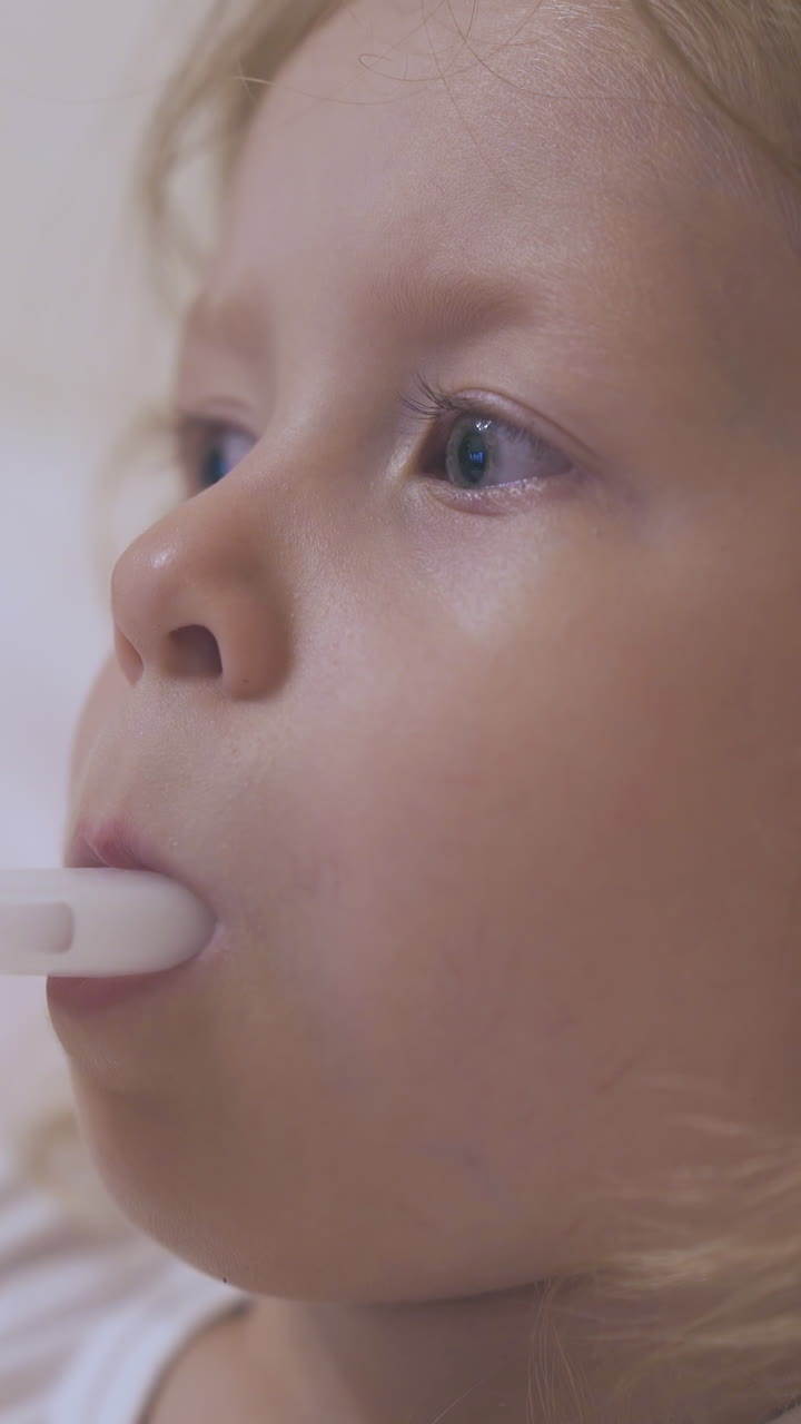 pretty blue eyed little girl breathes with modern inhaler lying in comfortable bed in light room extreme close view