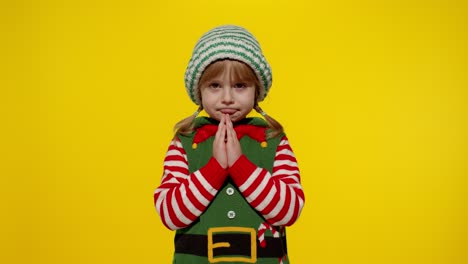 Kid-in-Christmas-elf-Santa-helper-costume-begs,-prays-about-gifts-on-New-Year.-Keeps-palms-together