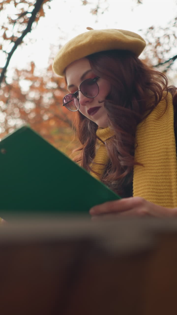dama elegante sentada centrada en su libro con una portada verde, rodeada de hojas secas de otoño con la luz del sol brillando a través, en un entorno de parque sereno