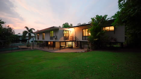 Impresionante-Toma-Al-Atardecer-De-Una-Gran-Casa-Elevada-De-Estilo-Tropical-Con-Césped-Verde-Y-Piscina-Y-Sorprendentes-Colores-De-Puesta-De-Sol-En-El-Fondo