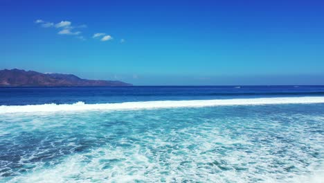 las olas blancas del océano azul profundo salpican hacia el fondo de la costa con altas montañas y un cielo azul brillante - tiro ancho